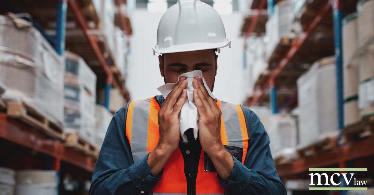 A warehouse worker wearing a hard hat blowing his nose in a warehouse