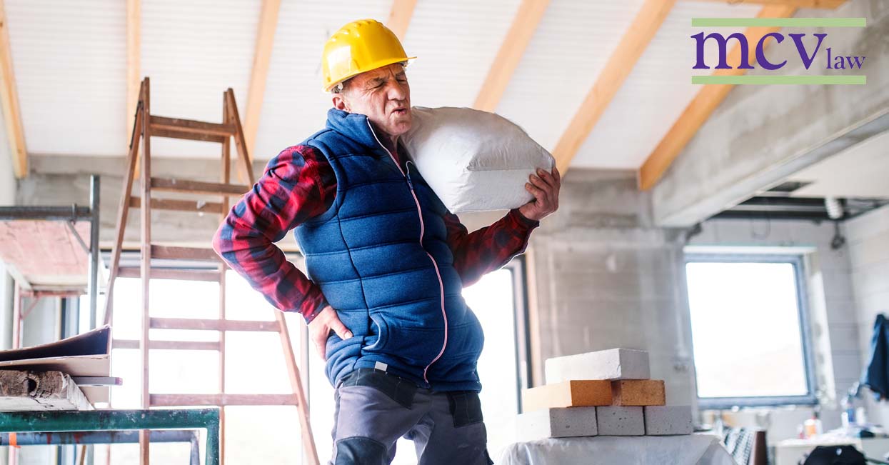A construction worker holding a heavy bag over his shoulder and holding his back in pain